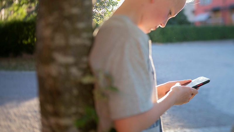 Barn står lutad mot ett träd och tittar ner i en mobiltelefon.