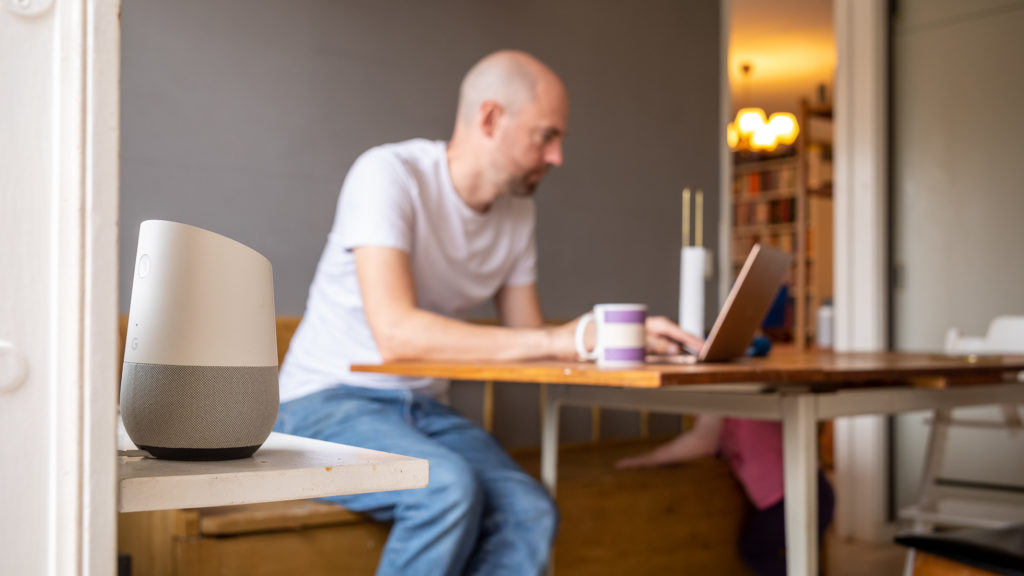 Man som sitter vid köksbordet och använder en dator, med en smart högtalare bredvid.