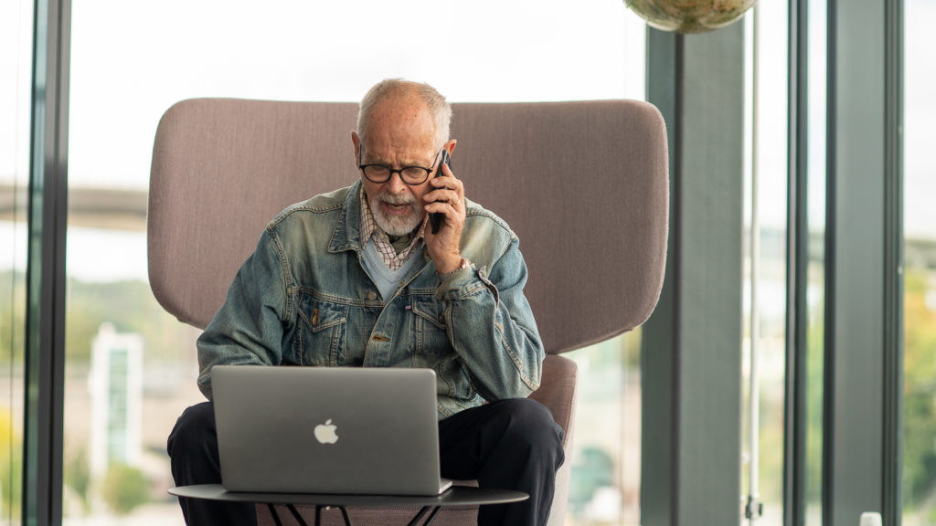 En äldre man med jeansjacka sitter i en fåtölj med en laptop framför sig och pratar i en mobiltelefon