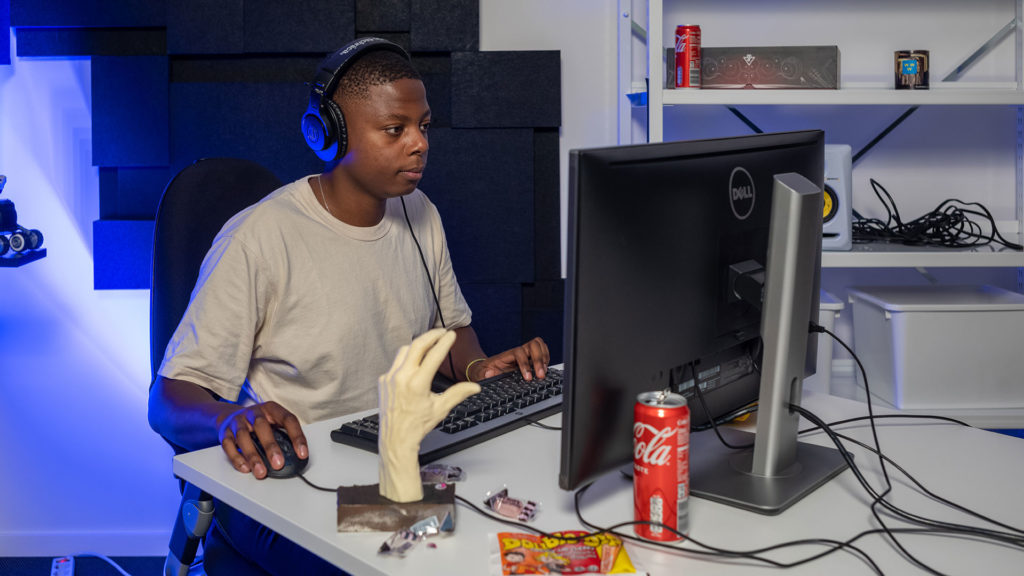 En kille sitter med hörlurar, tangentbord och mus framför en datorskärm på ett skrivbord