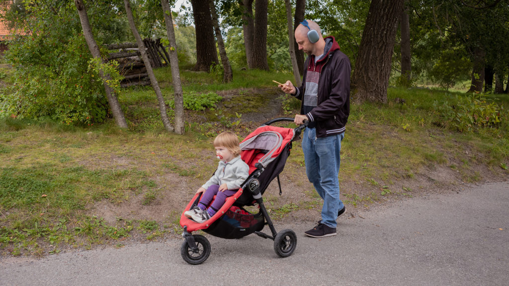 En pappa väljer något att lyssna på i sin mobil samtidigt som han går med sitt barn i vagnen.