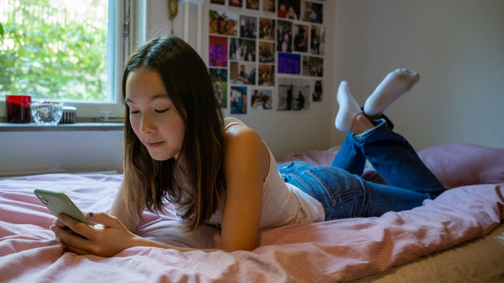 Ung tjej ligger på sin säng och surfar på sin smartphone.