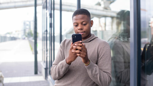 En ung kille står lutad mot en vägg och tittar på sin mobil.