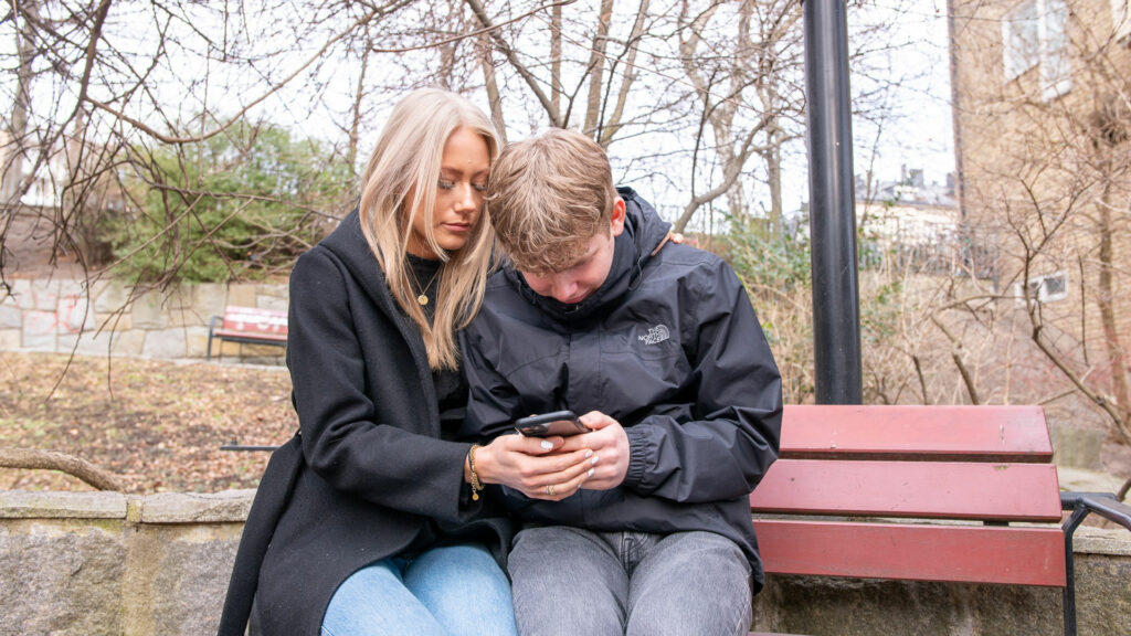 En ung tjej håller om en ung kille som ser ledsen ut när han tittar på sin mobil. De sitter på en parkbänk.