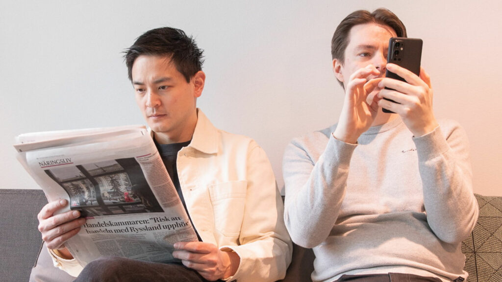Två män sitter i en soffa, den ena läser papperstidning, den andra kollar på mobilen.