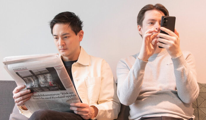 Två män sitter i en soffa, den ena läser papperstidning, den andra kollar på mobilen.