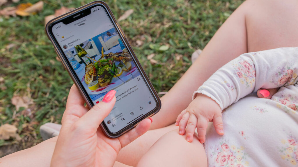 Instagram i Sverige. En hand som håller en mobil, på skärmen syns Instagram-flödet. Andra handen hålls om en bebis.