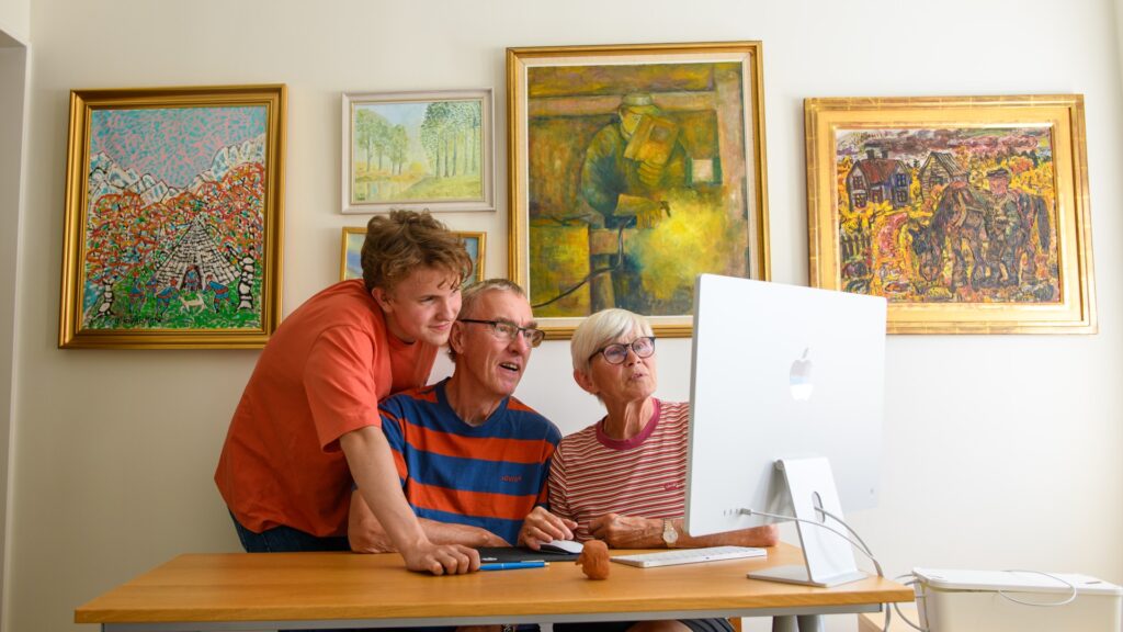 En ung man står bredvid ett skrivbord där ett äldre par sitter framför en dator, han ser ut att hjälpa paret.