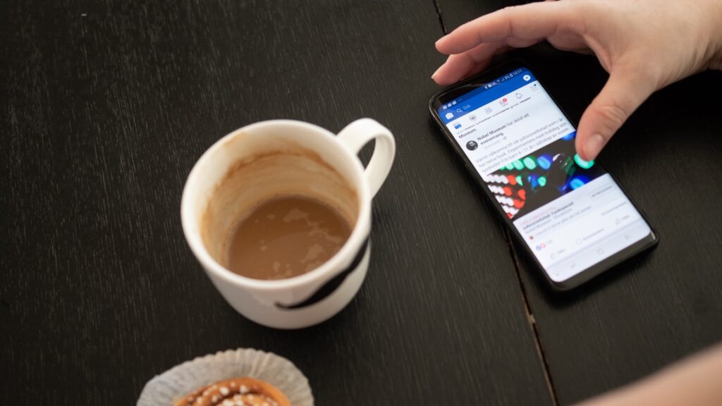 Händer som scrollar på en mobil vid ett bord, på bordet står en kaffekopp och en bulle. Facebook syns på mobilens skärm. 