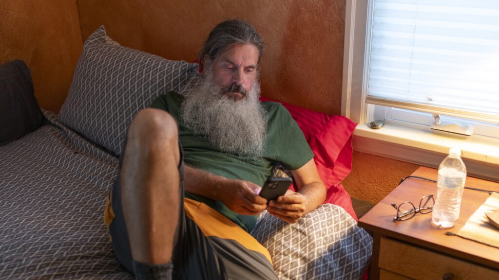 En man tittar på sin mobil, han ligger på sängen med ena benet uppdraget, på nattygsbordet ligger ett par glasögon.
