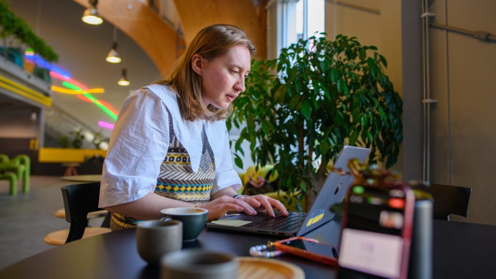 En kvinna sitter på ett café och jobbar på sin dator, i bakgrunden syns neonlampor.