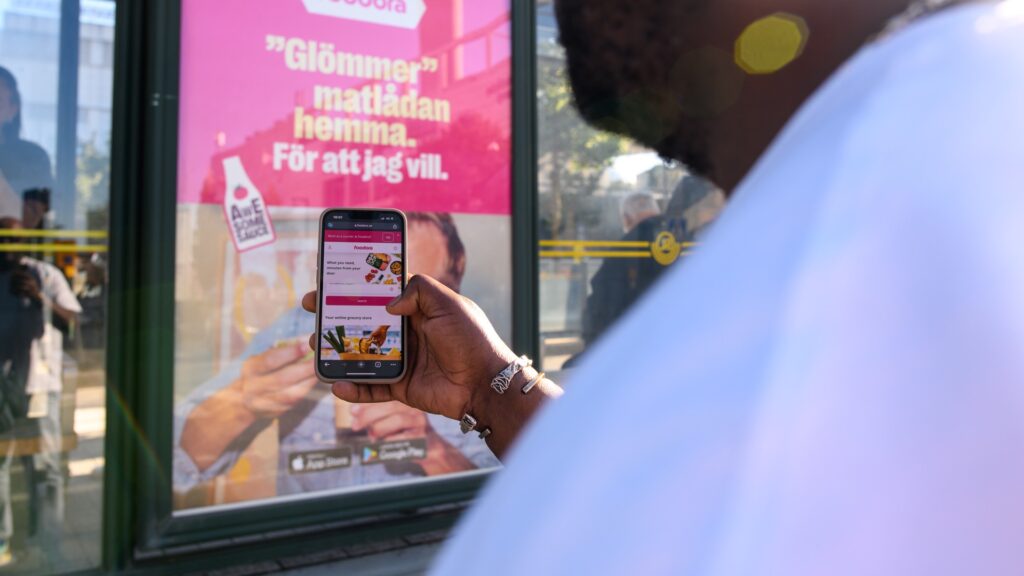 En man går förbi en reklamskylt med Foodora på, i hans mobil syns samma reklam på skärmen – riktad reklam.