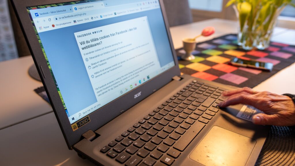 En dator på ett köksbord, på skärmen syns en text om cookies. En äldre persons hand trycker på tangenterna.