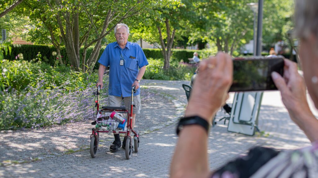En äldre man går med rullator, i förgrunden syns en äldre kvinna som tar en bild på mannen med mobilkamera.