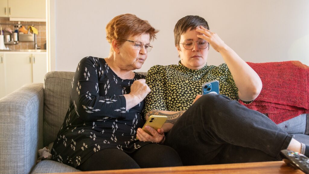 Två kvinnor sitter i en soffa och tittar på sina mobiler, de ser oroliga ut. Den ena tar sig för pannan.