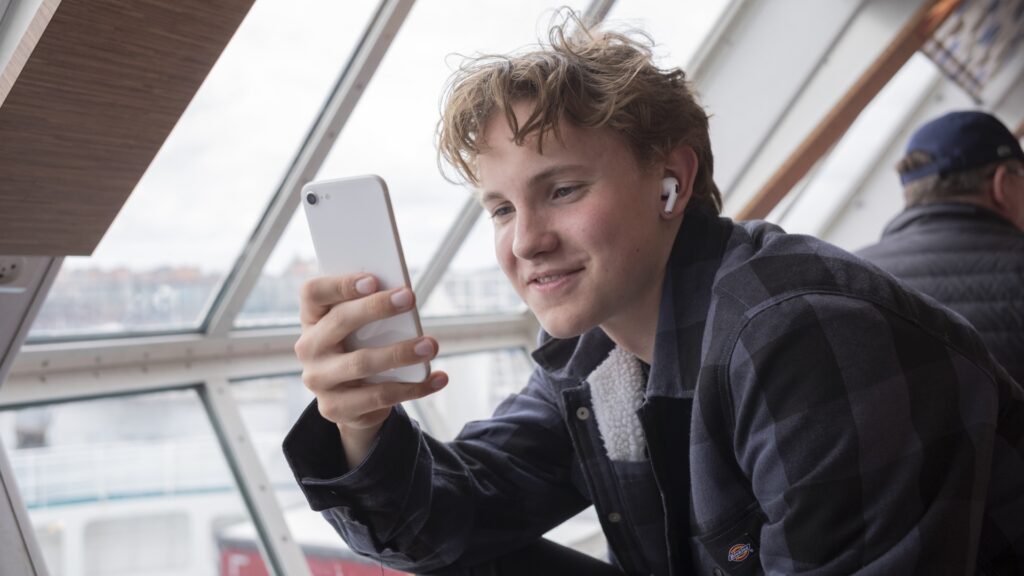 En ung man sitter vid ett fönster och tittar på sin mobil, han har airpods i öronen och ser glad ut.