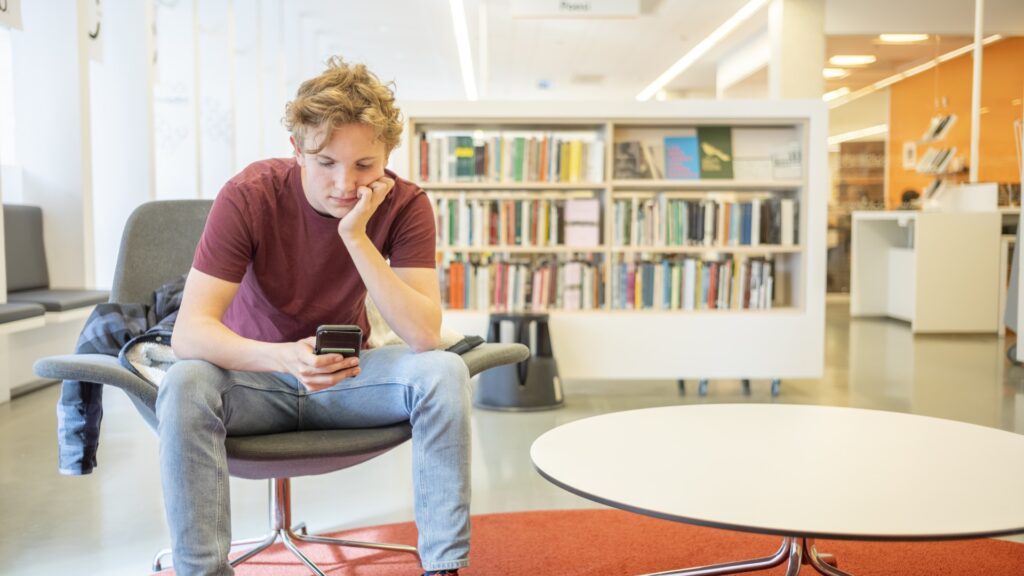 En ung man sitter i en fåtölj på ett bibliotek, han håller sin mobil i handen, i bakgrunden syns böcker.