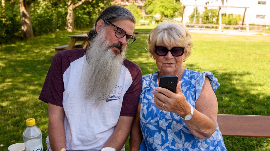 Två äldre personer sitter utomhus, de tittar tillsammans på en mobil.
