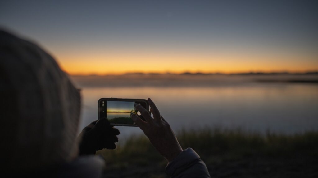 En person håller upp sin mobil och tar en bild av en vacker solnedgång. Hållbarhet, klimat, internet i Sverige.