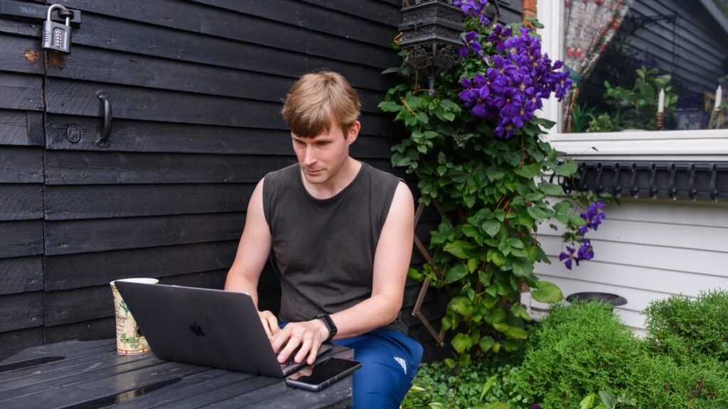 En man sitter utomhus med en laptop framför sig på ett trädgårdsbord.