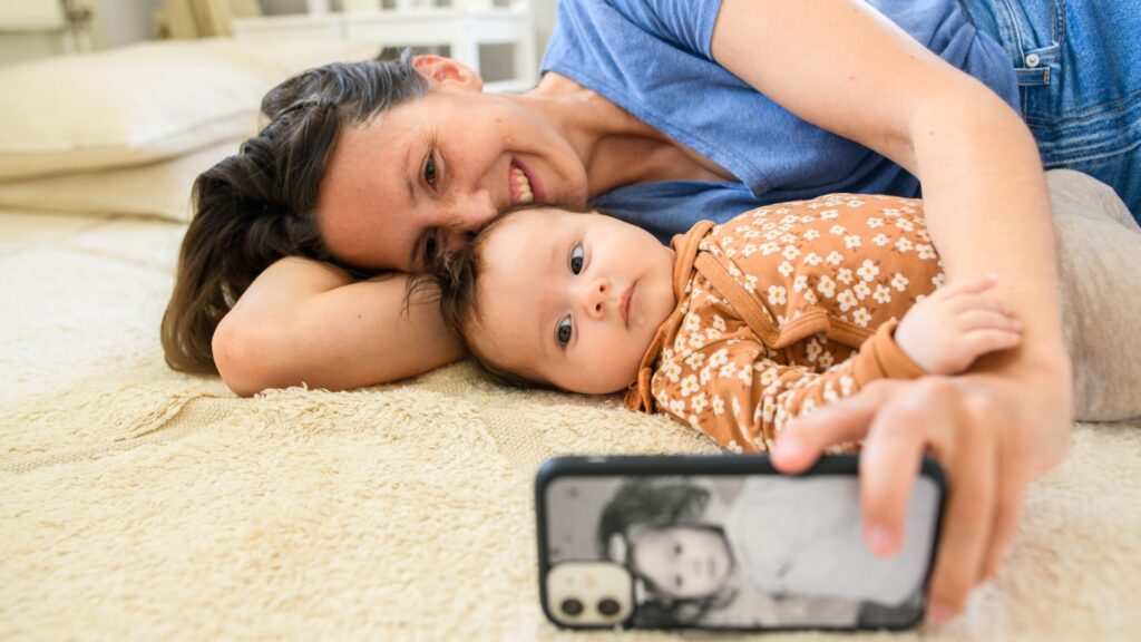 En kvinna ligger på mattan med sin bebis, hon tar en selfie till sociala medier.