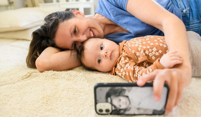 En kvinna ligger på mattan med sin bebis, hon tar en selfie till sociala medier.