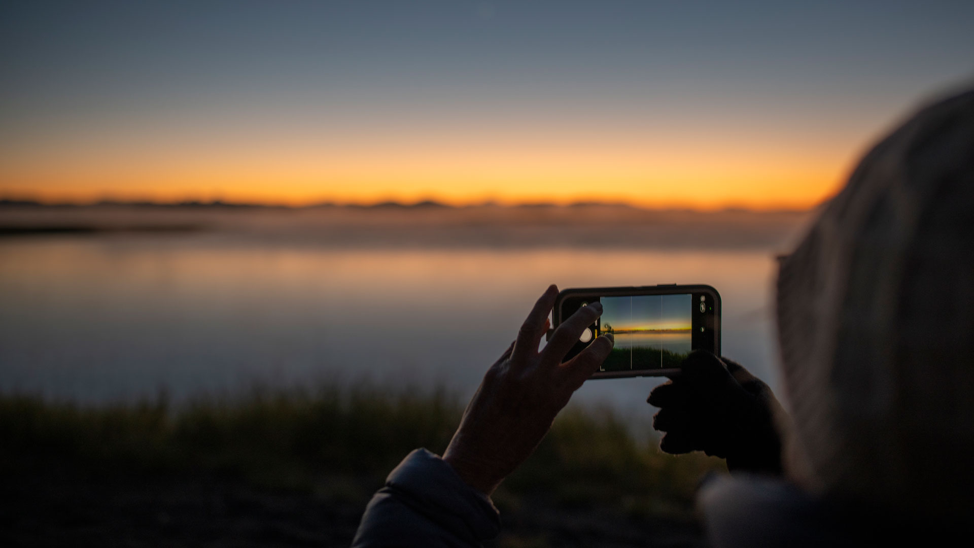 En person håller upp en mobilkamera mot en solnedgång.