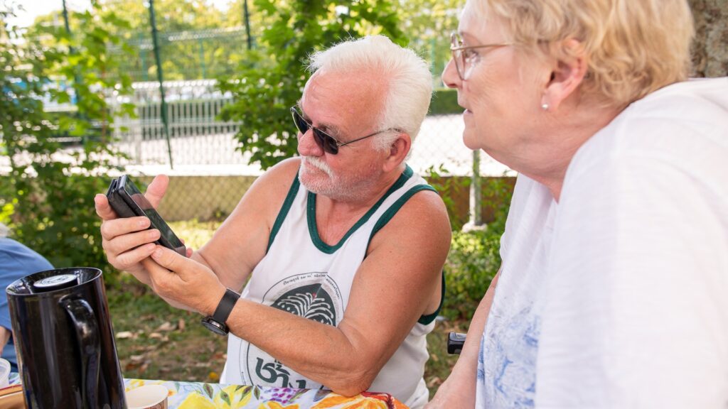 En äldre man iklädd linne visar mobilen för en äldre kvinna, de är utomhus och det är sommar.