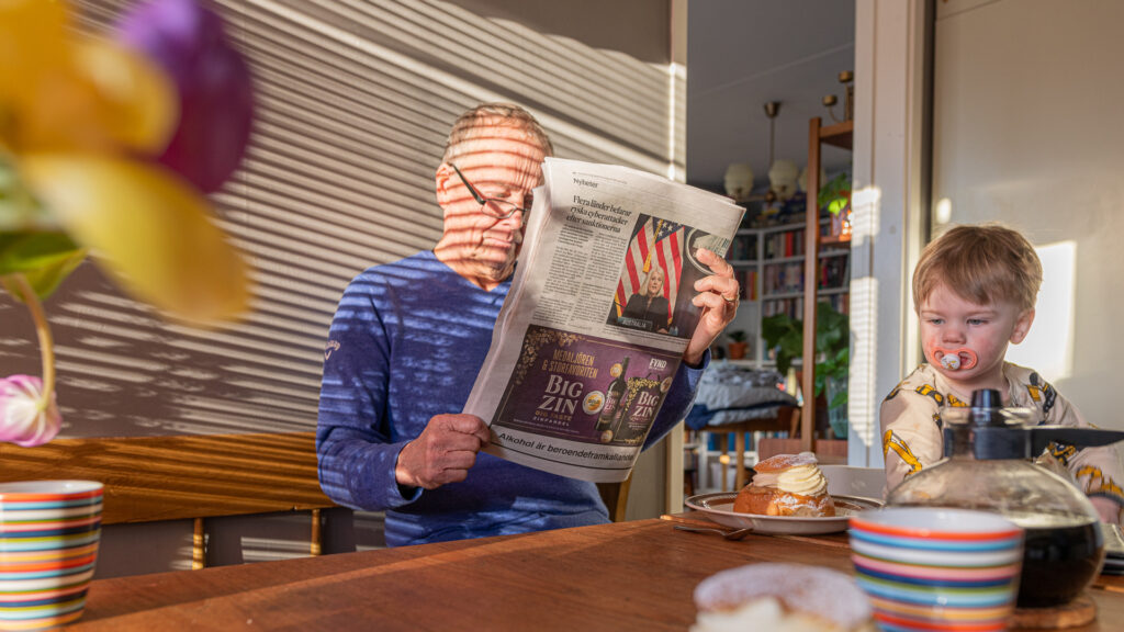 En man sitter vid ett köksbord tillsammans med ett barn. Mannen läser en papperstiding.