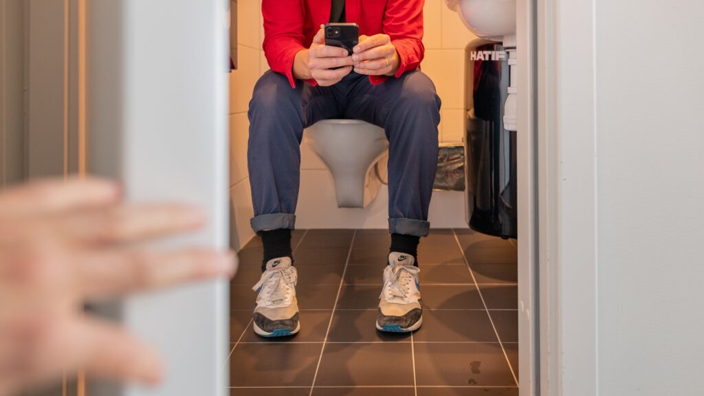 En kille sitter på toaletten och scrollar på sin mobil.