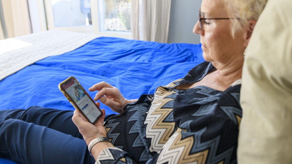 En äldre kvinna sitter i sängen och loggar in på 1177 på mobilen.