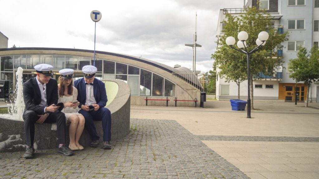 Tre ungdomar med studentmössa sitter och scrollar på mobiler.