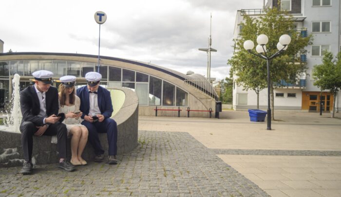 Tre ungdomar med studentmössa sitter och scrollar på mobiler.