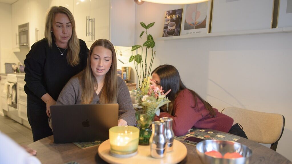 En förälder står vid sidan av bordet där hennes barn tittar på skärm.
