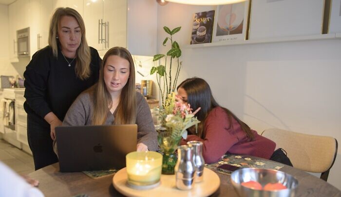 En förälder står vid sidan av bordet där hennes barn tittar på skärm.