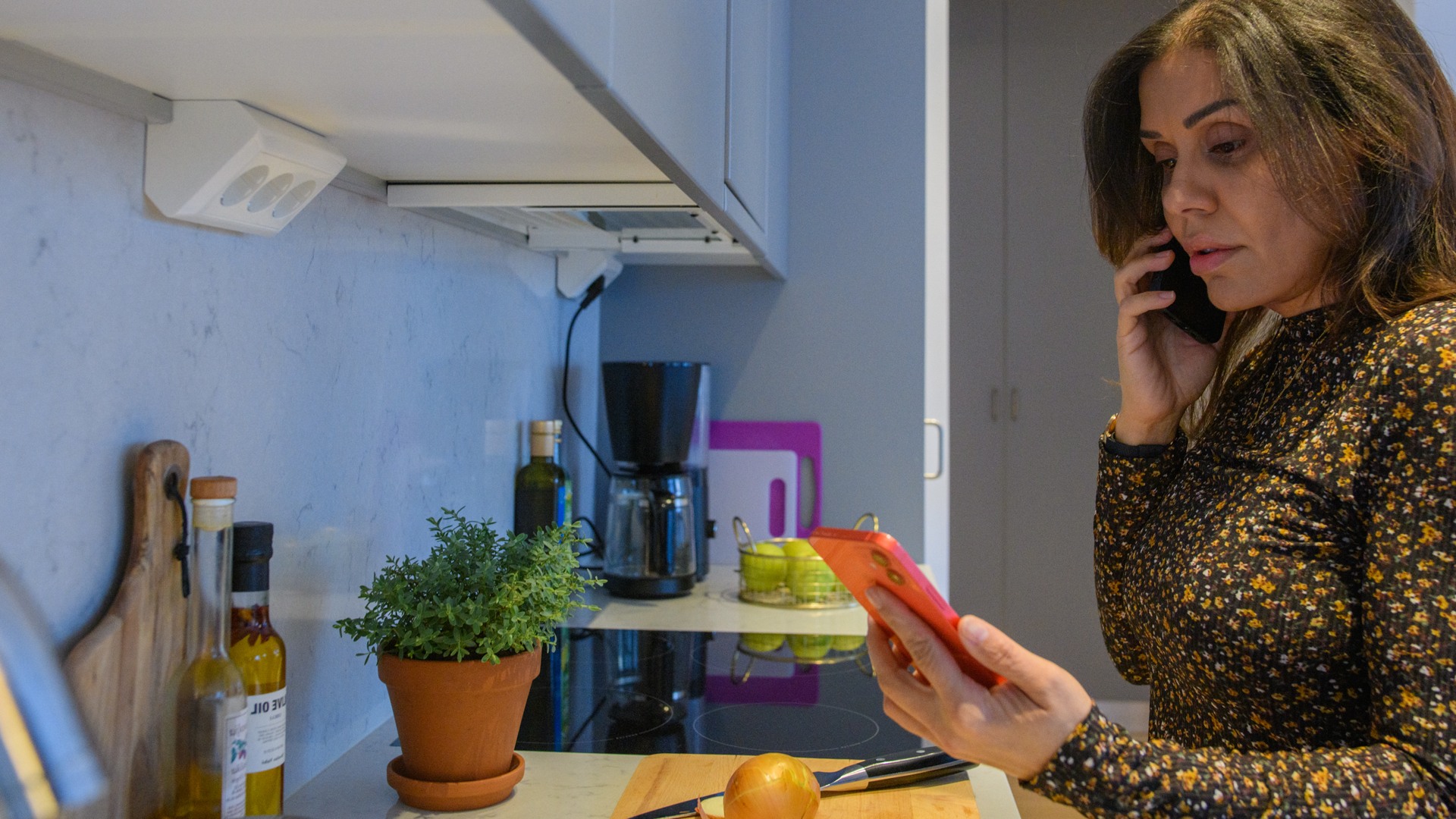 En kvinna i ett kök som pratar i mobiltelefon samtidigt som hon kollar partnerns mobil.
