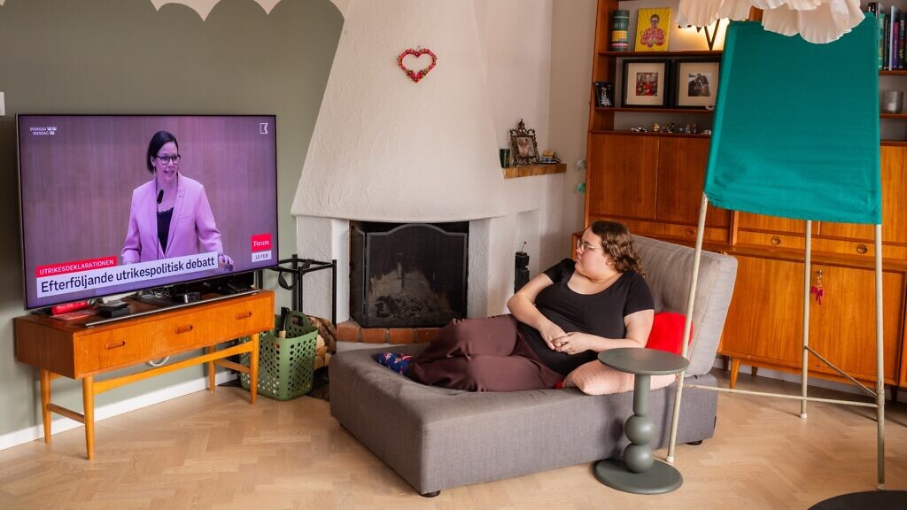 En kvinna sitter i en stol framför en tv och tittar på ett politiskt inslag på Kunskapskanalen.
