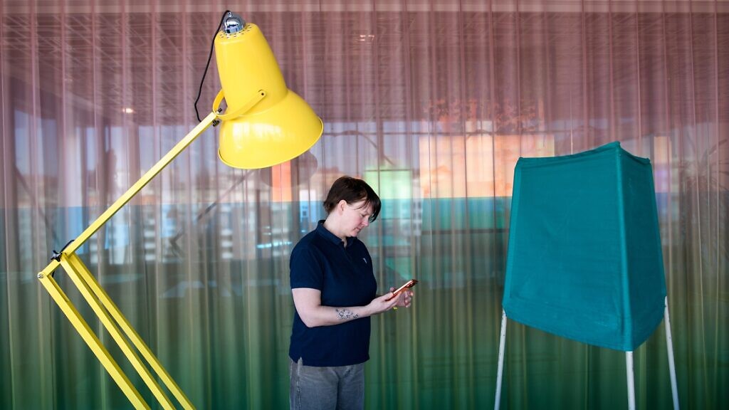 En kvinna står mellan en gul lampa och en valskärm och tittar på sin mobil.