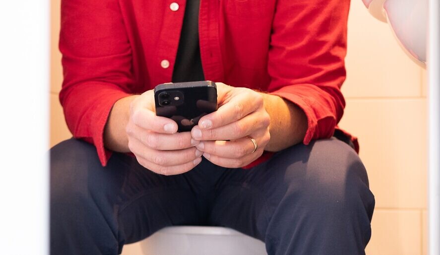 En man sitter på en toalett och håller i en mobiltelefon.