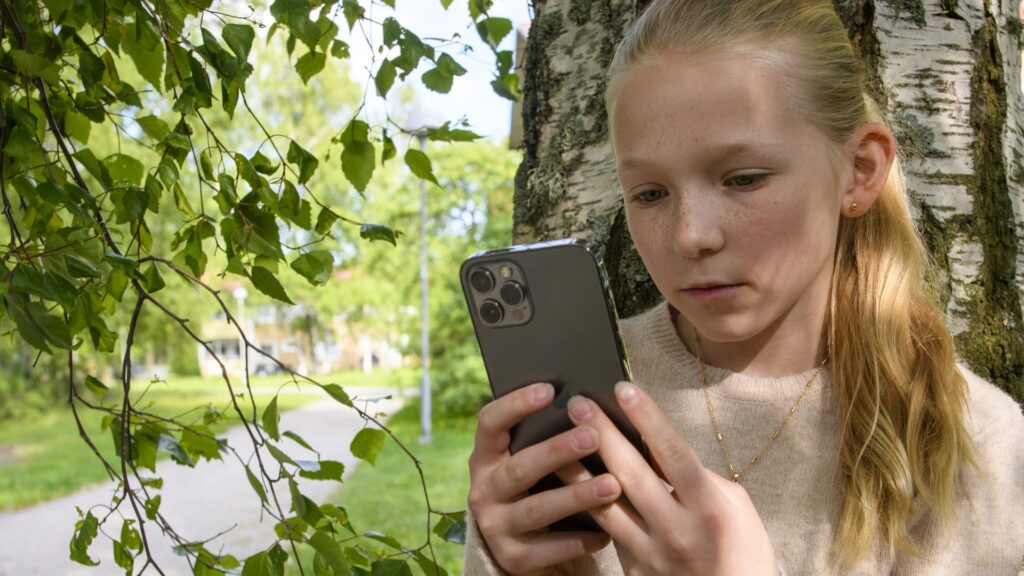 En flicka står lutad mot ett träd och scrollar på sin mobil.