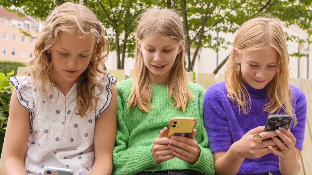 Tre flickor sitter på en bänk och tittar på sina mobiltelefoner.